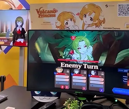 A Volcano Princess display at a shared booth, with the game running on a monitor beneath the banner.